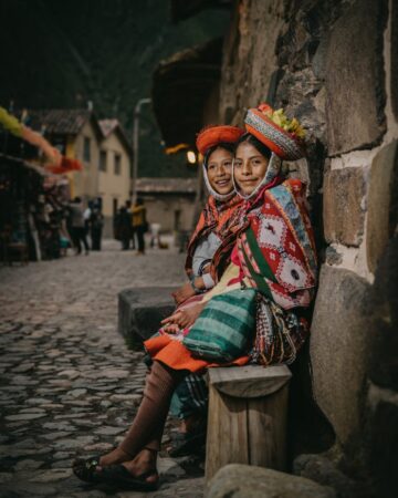 Two women in traditional attire seated on cobblestones in Ollantaytambo, Peru.